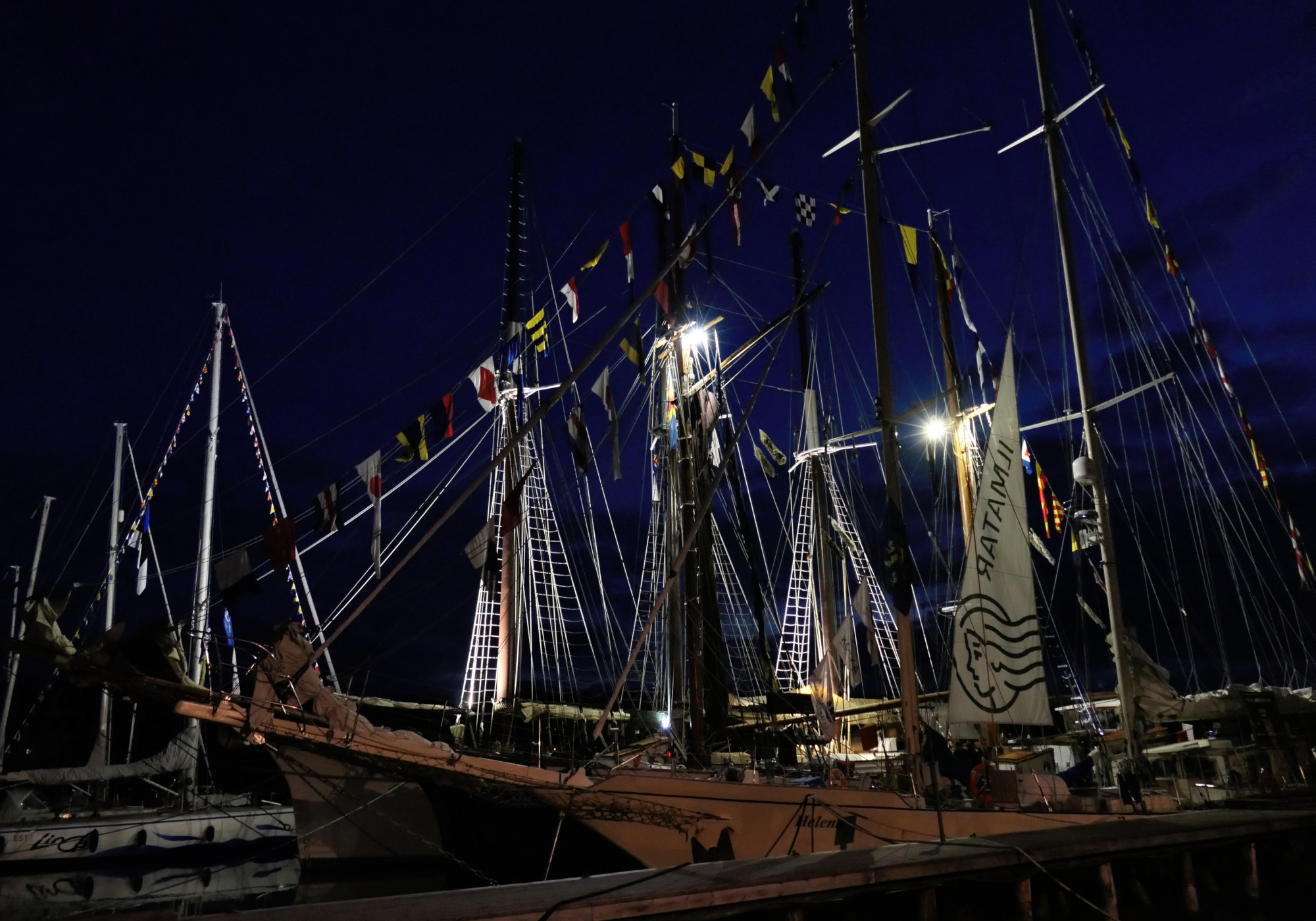 Skepp i hamn i nattljuset med belysning bakifrån så att masterna lyses upp. I bakgrunden syns några enstaka master från segelbåtar i hamn.