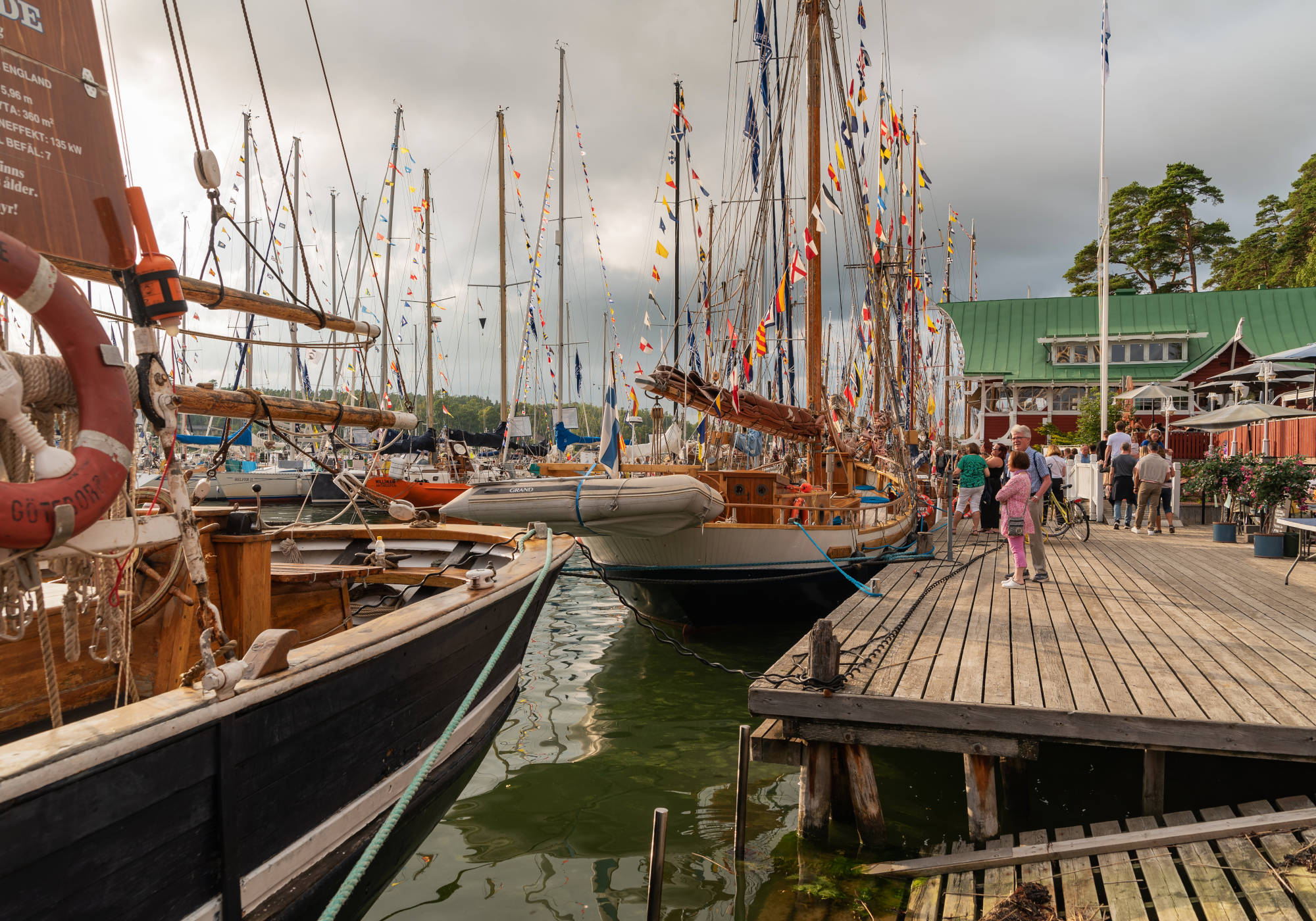 Skepp och segelbåtar ligger sida vid sida i hamn med människor på en terass som betraktar båtarna. I slutet av terassen syns en byggnad och dess uteservering.