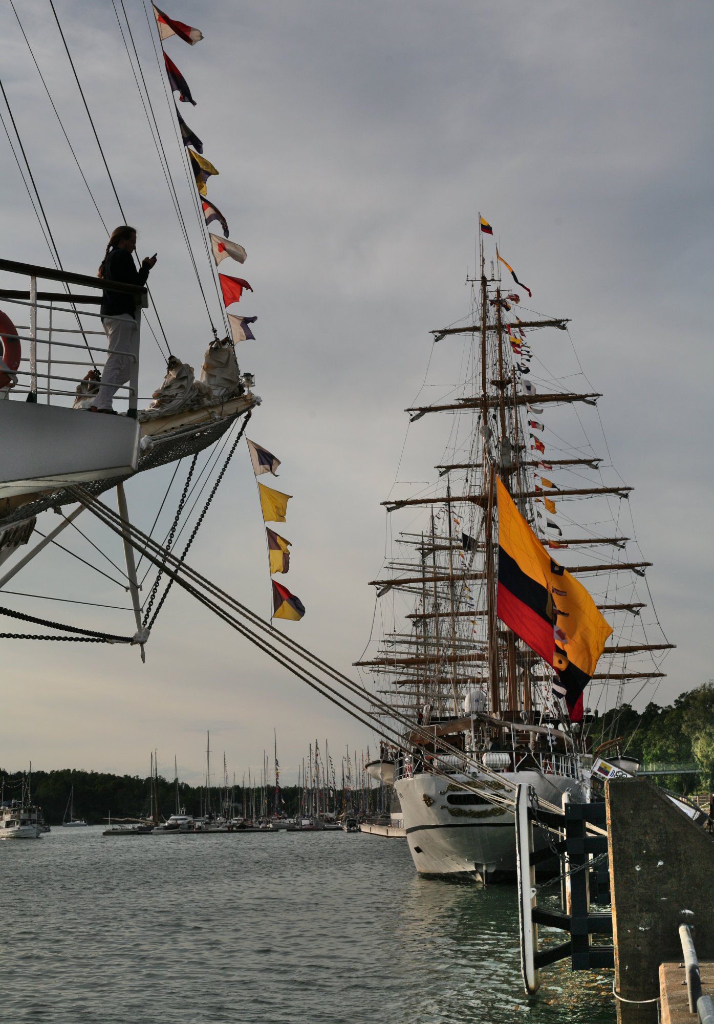 En person ses i fören på ett fartyg, spejandes mot ett annat skepp som ligger i hamn. I bakgrunden syns många segelbåtar med deras master förtöjda vid bryggor.