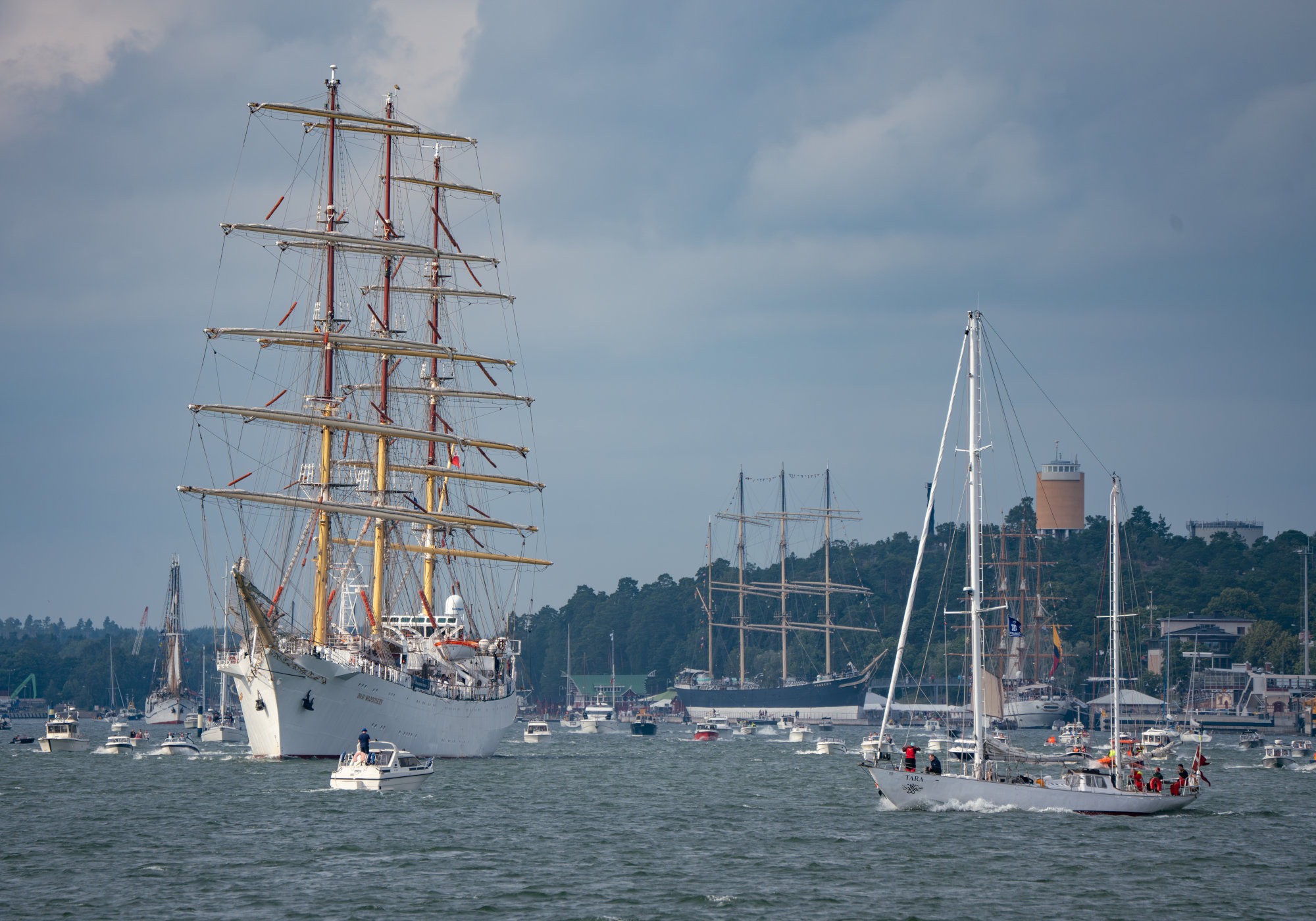 Skeppet Dar Mlodziezy är på väg ut från hamnen i Mariehamn. Runtom ses många småbåtar och en större segelbåt. I bakgrunden syns segelfartyget Pommern, vattentornet i Mariehamn och skog.