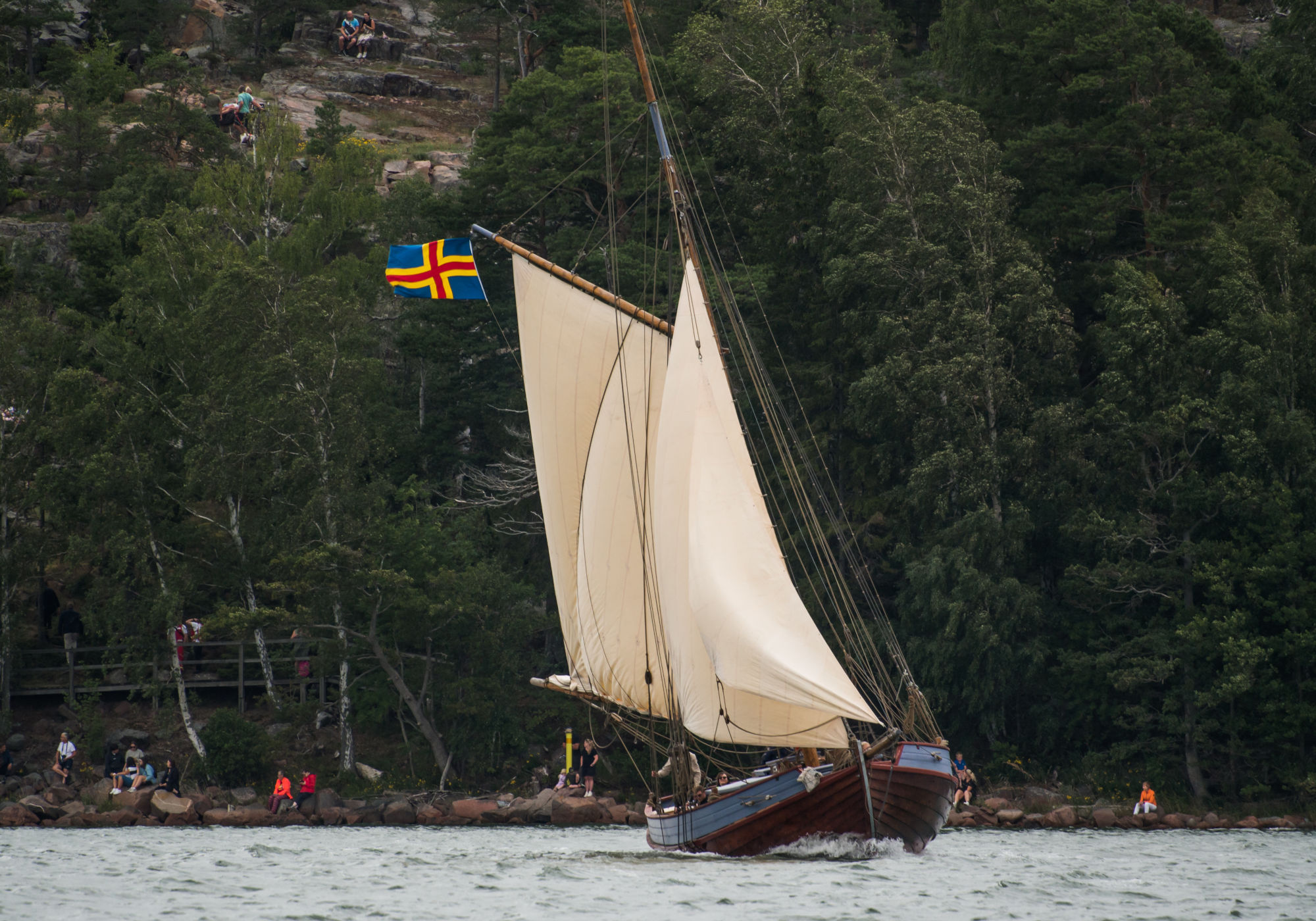 Alanta ses med fulla segel på kryss utanför Mariehamn med Ålands flagga som fladdrar i vinden. I bakgrunden syns människor längs strandlinjen och uppe på klipporna i skogen.