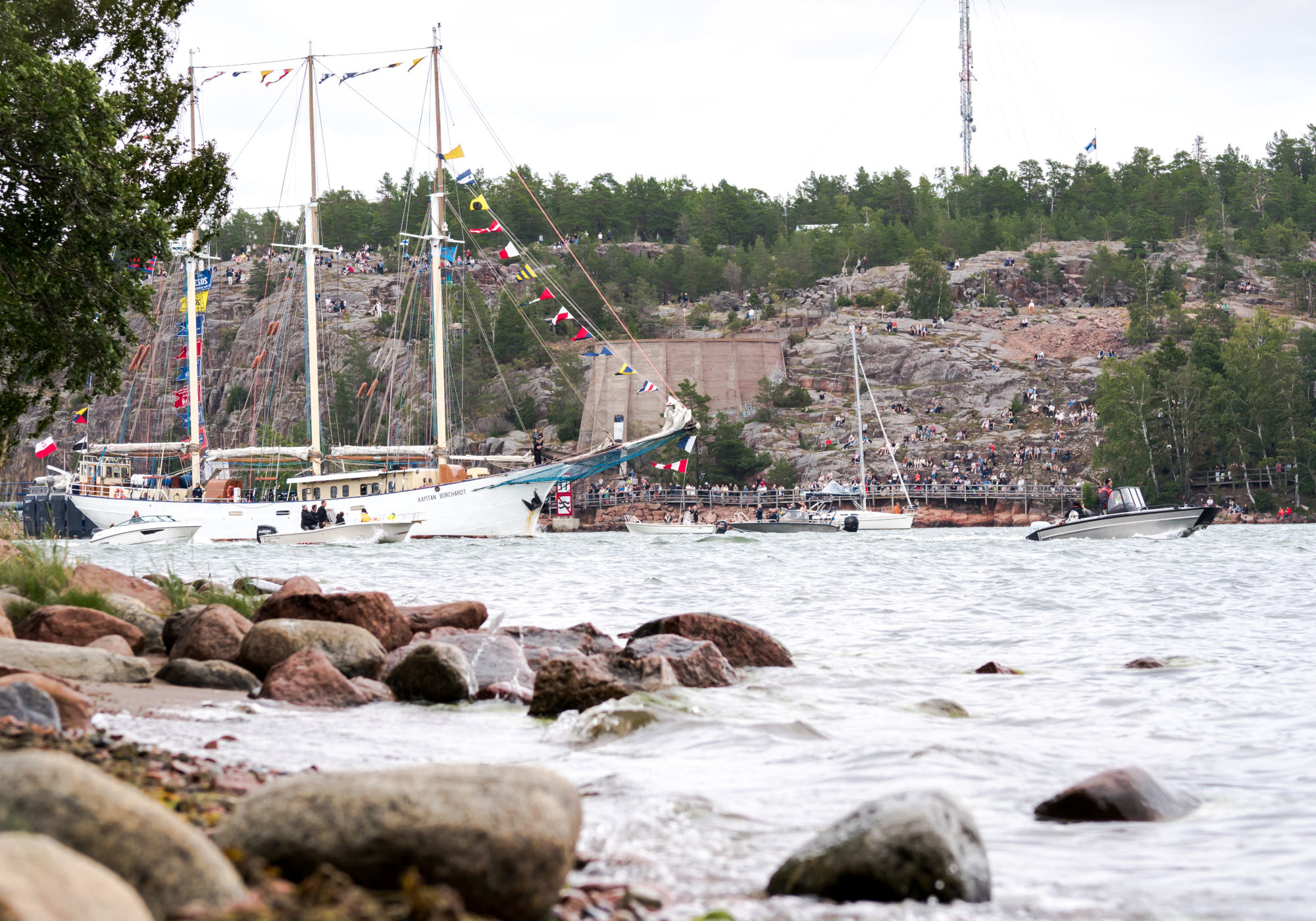 Skeppet Kapitan Borchardt har vimplar i masterna. Runt om ses mindre motorbåtar och en segelbåt. I förgrunden syns stenar vid strandkanten och i bakgrunden ses människor längs med strandpromenaden och uppe i bergen ovanför.
