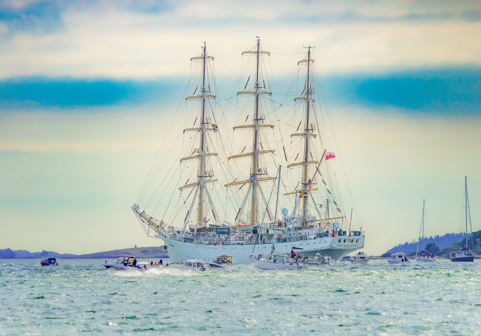 Skeppet Dar Mlodziezy lämnar Mariehamn på sin väg ut mot havet med många motorbåtar och segelbåtar runt om. Den polska flaggan vaggar i aktern och i en mast fladdrar Ålands flagga. I bakgrunden syns öar och skär.